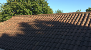 "Photorealistic image focusing on a section of a typical UK residential tiled roof. A clear, distinct shadow from a large nearby tree (the tree itself can be partially out of frame) falls across a significant portion of the roof where solar panels could be installed, visually illustrating the impact of shading. Bright sunshine highlights the contrast between the lit and shaded areas."